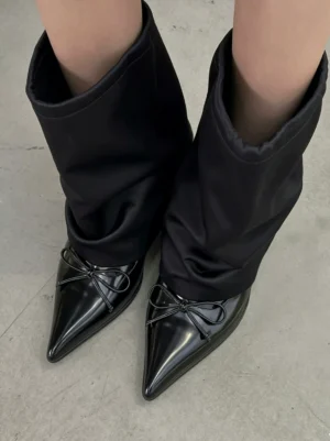 Top view of black satin slouch pointed-toe mid-calf boots with patent-look bow toes worn on concrete floor