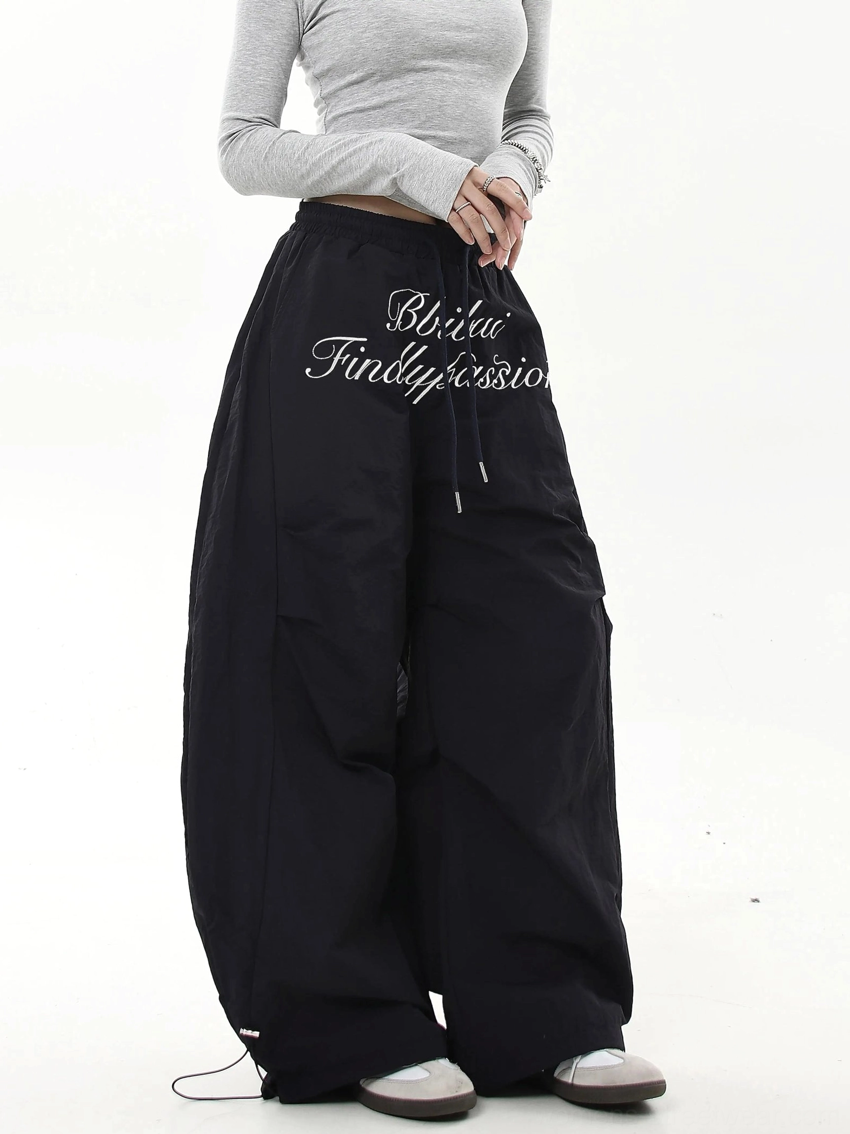Black oversized parachute streetwear cargo pants shown from a side-front angle with white script embroidery and exaggerated wide legs