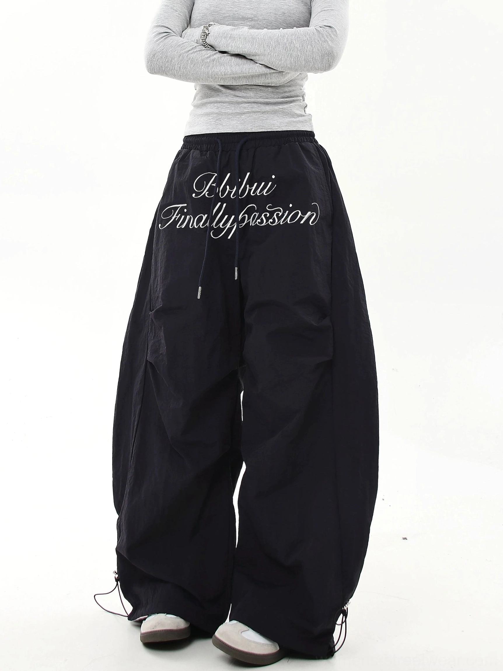 Back view of black oversized parachute cargo pants with white script embroidery, wide baggy legs and adjustable bungee ankle cuffs