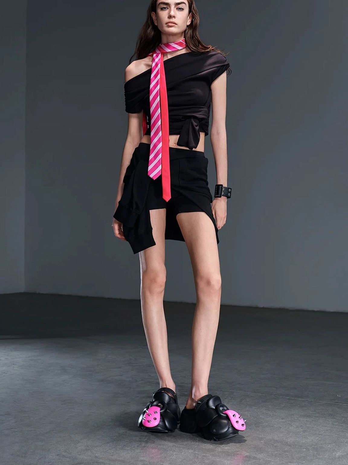Side styling view of black sculpted platform shoes with pink floral spike details worn with a black mini skirt and striped tie