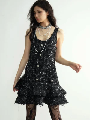Front three-quarter view of a black sequin tweed tiered mini dress styled with layered necklaces, fishnet tights, kitten heels, and a compact clutch