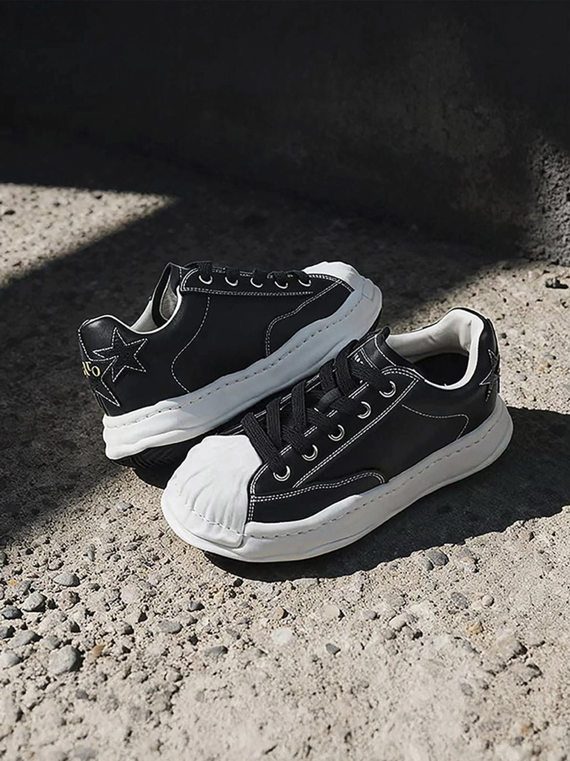 Black shell-toe court sneakers on concrete in a dim street setting, showing white topstitching and contrasting dissolving-style sole