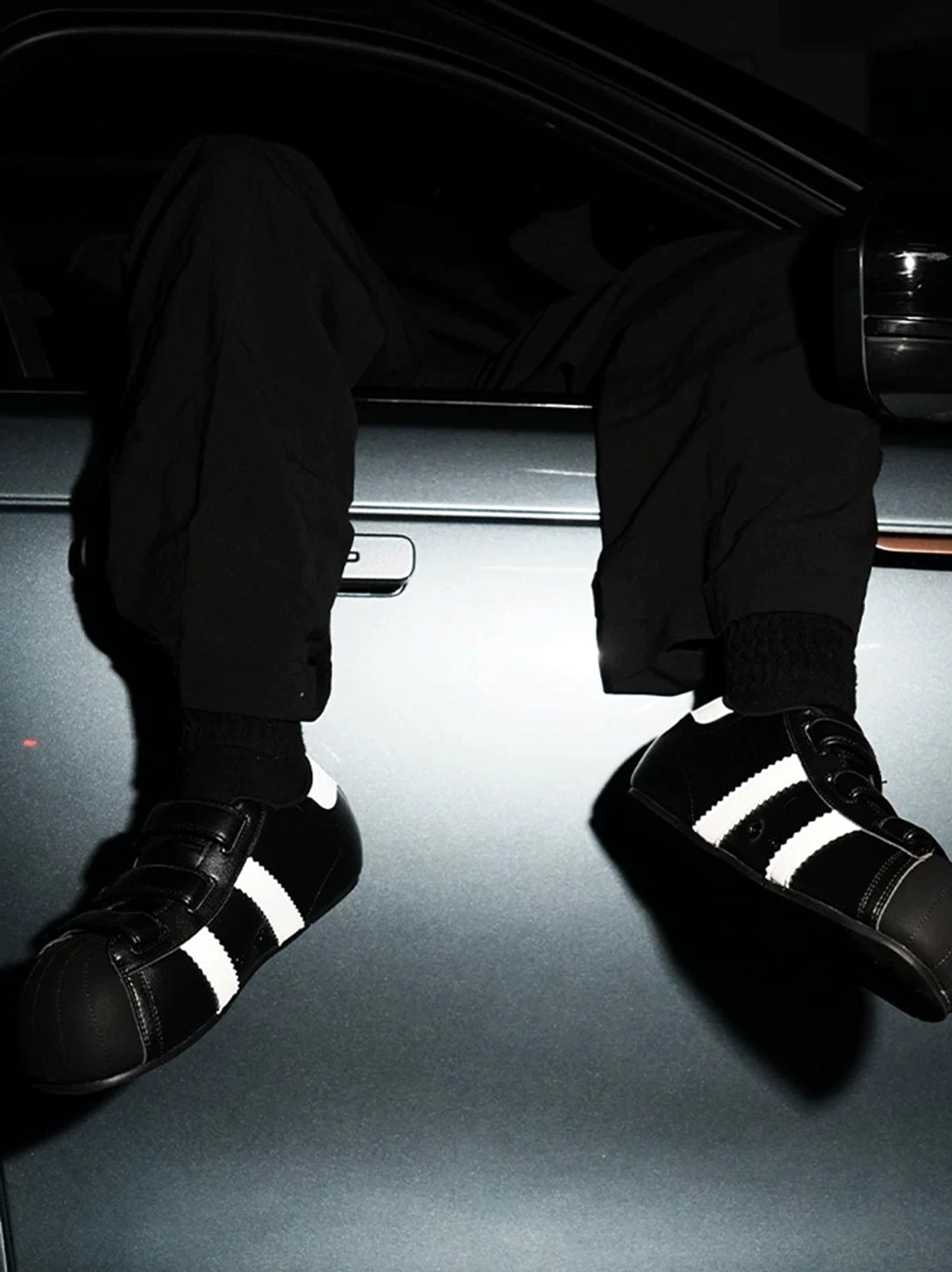 Black shell toe hidden lift sneakers styled with all-black streetwear pants beside a car