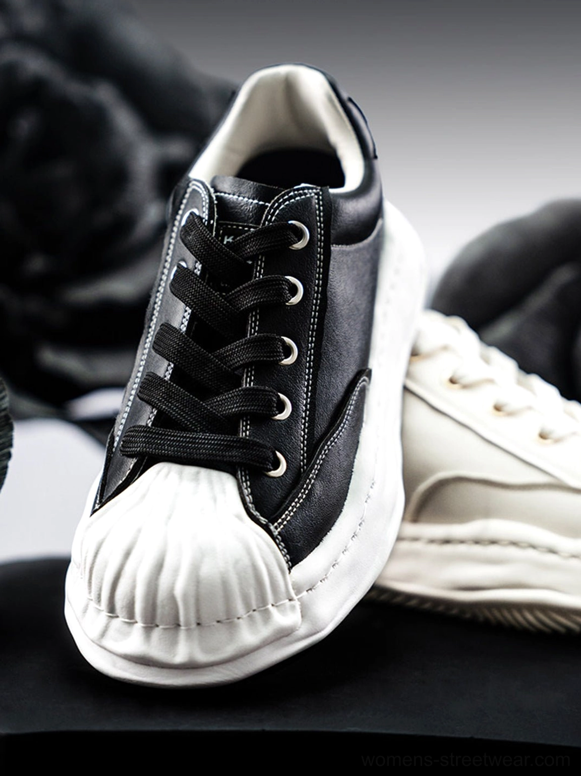 Extreme close-up of black shell-toe sneaker showing textured molded toe, metal-look lace eyelets, and black flat laces