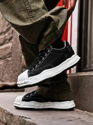 Black shell-toe streetwear sneakers worn with olive green cargo pants on outdoor stairs, showing contrast stitching and a thick white court outsole