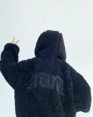 Back view of black oversized sherpa zip hoodie showing gothic lettering, plush teddy fleece texture, attached hood, and relaxed streetwear fit