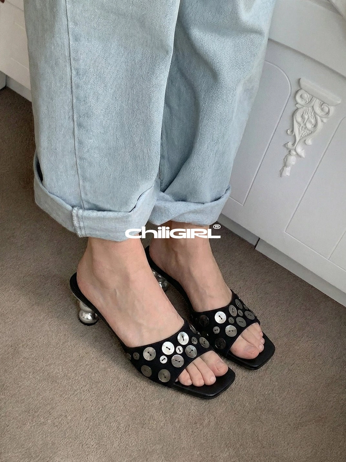 Black sculpted heel slide sandals with silver circular embellishments styled with rolled light wash jeans