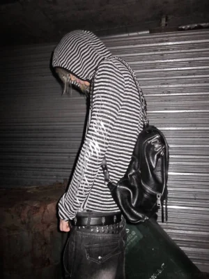 Back view of a black and silver vertical striped hooded cover-up shirt styled with a leather backpack and dark urban streetwear outfit