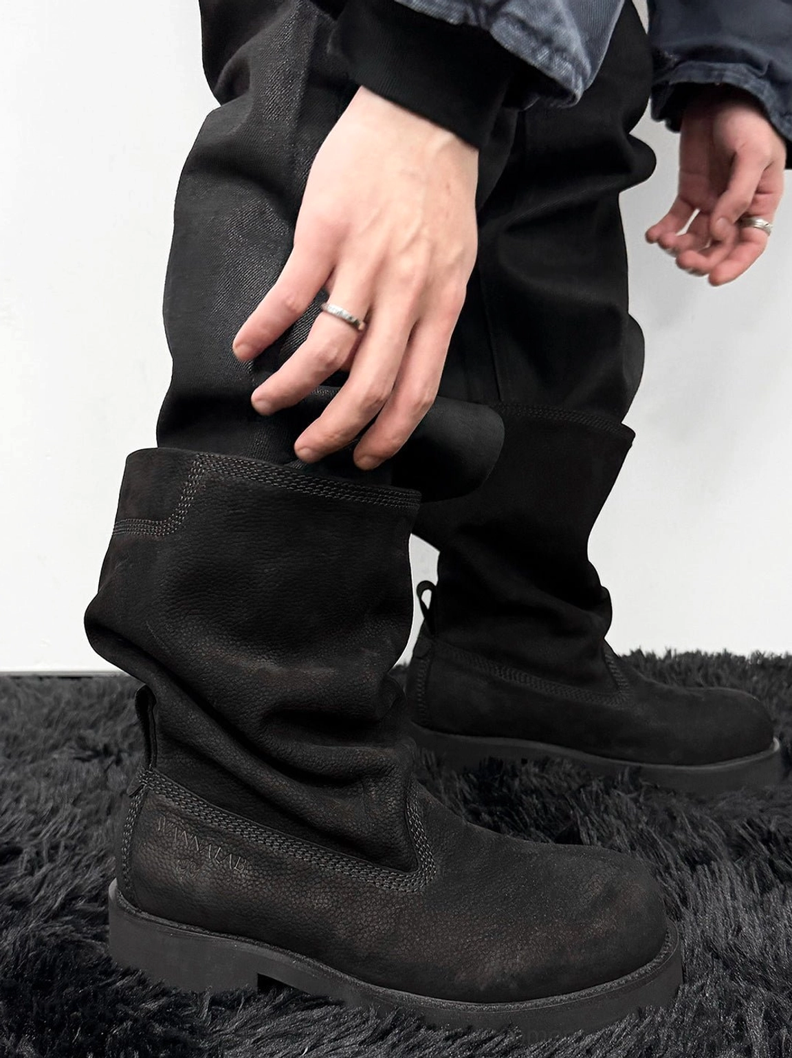Rear side view of black slouch mid-calf streetwear boots worn with dark clothing, highlighting the relaxed shaft and rugged chunky rubber sole