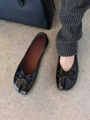 Black split-toe leather ballet flats with bow detail styled on feet with leather handbag on concrete floor
