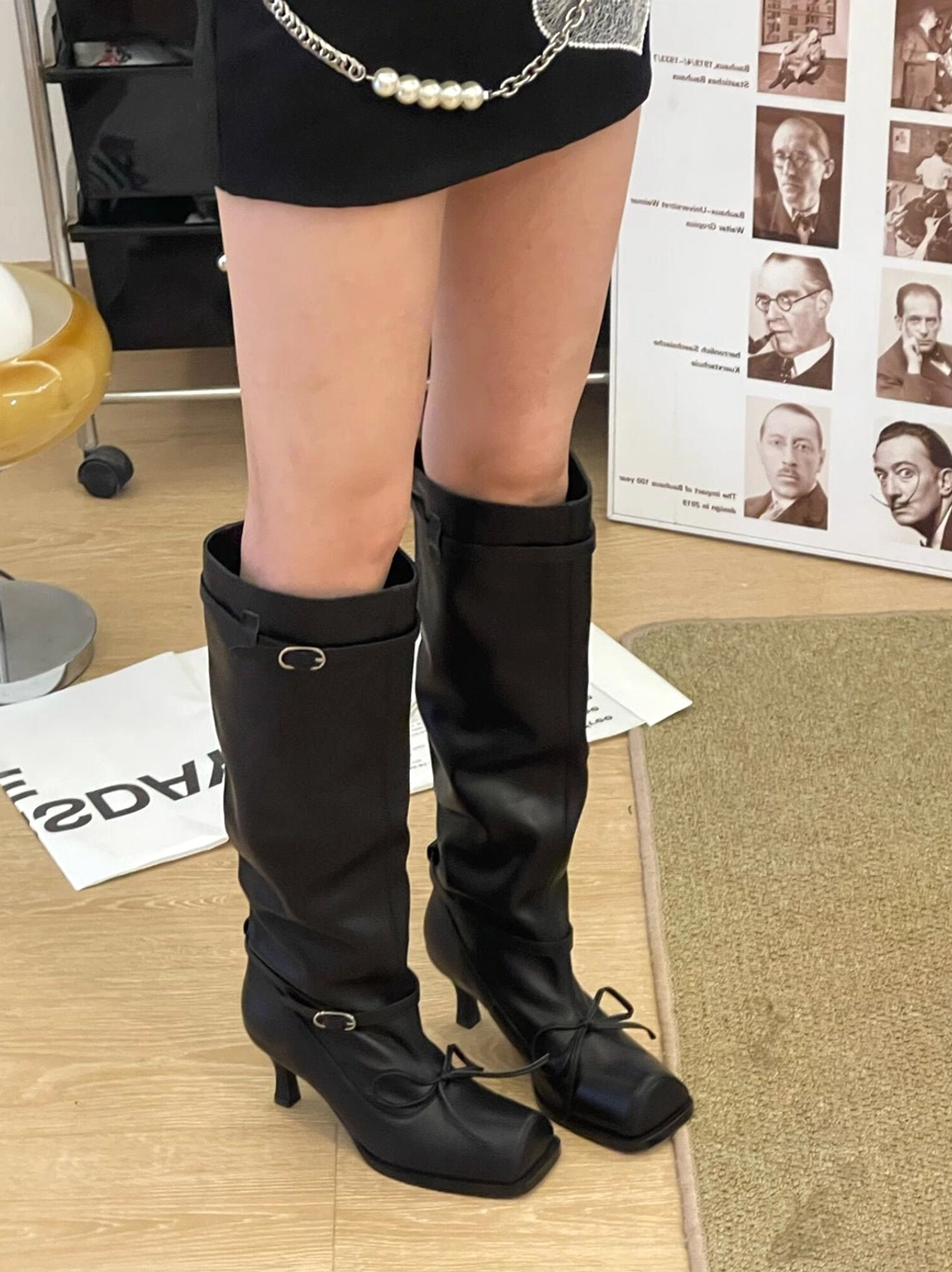 Front three-quarter view of black square-toe knee-high leather boots with bow-accented strap, relaxed shaft, metal buckle, and low curved heel