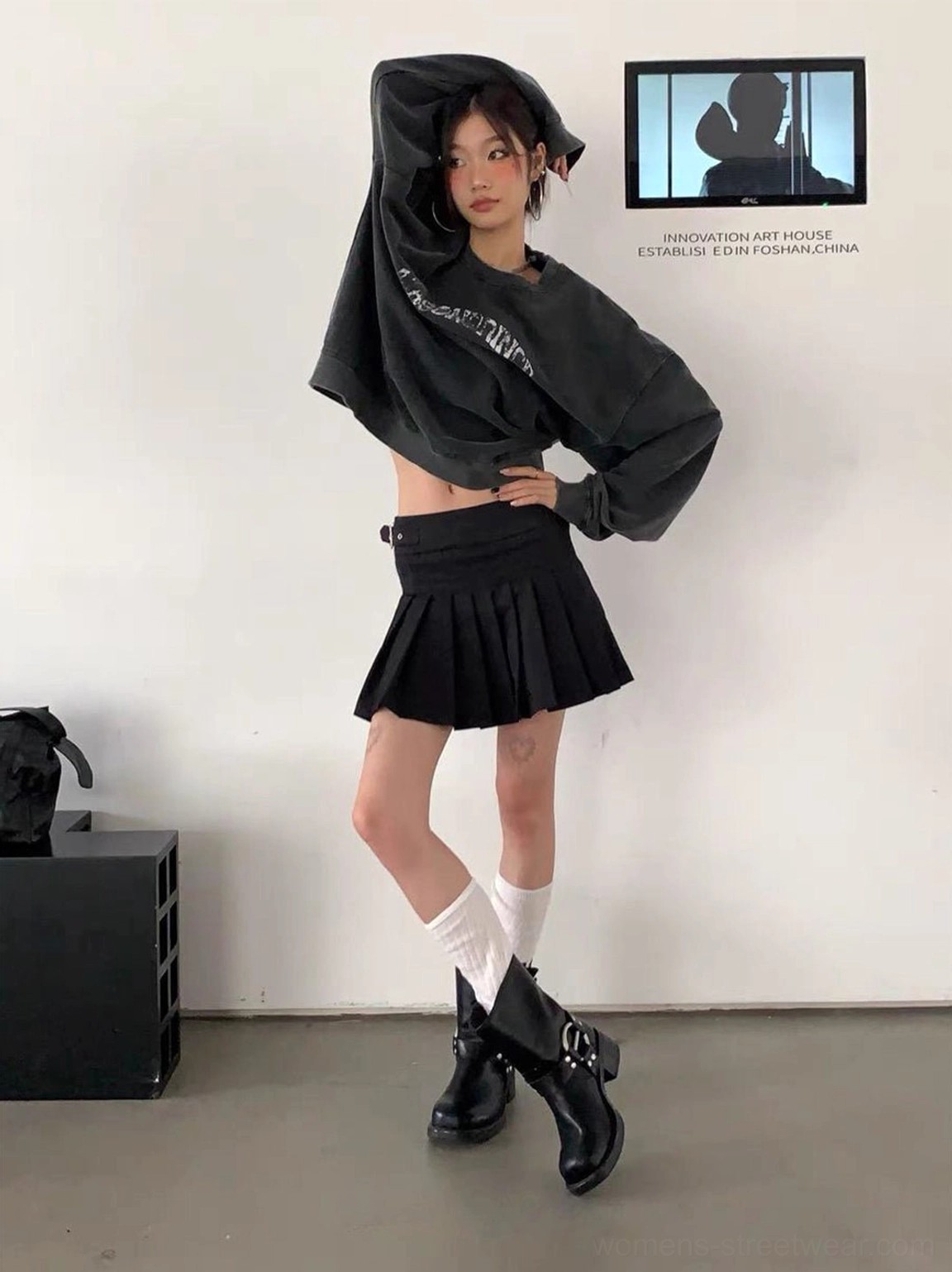 Woman wearing black square-toe leather Western boots with buckles, cropped sweatshirt, pleated mini skirt, and white socks