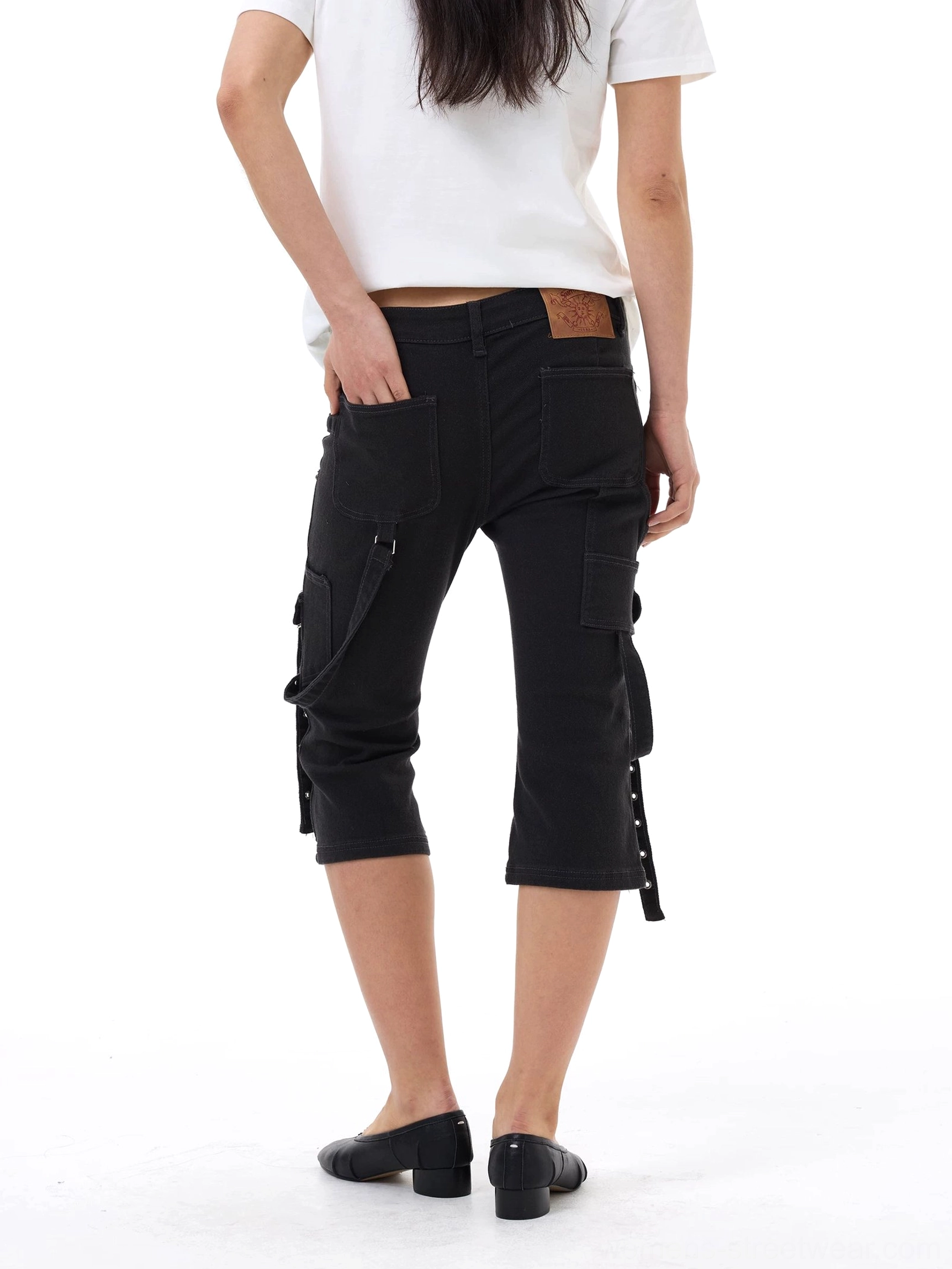 Back view of black strap cargo capri jeans with a back waist patch, slim cropped silhouette, side cargo pockets, and hanging utility straps