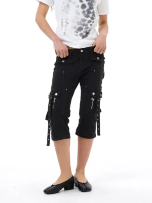 Front view of black strap cargo capri jeans styled with a printed white T-shirt and black flats, showing slim cropped fit, utility zipper pockets, metal rivets, and hanging side straps