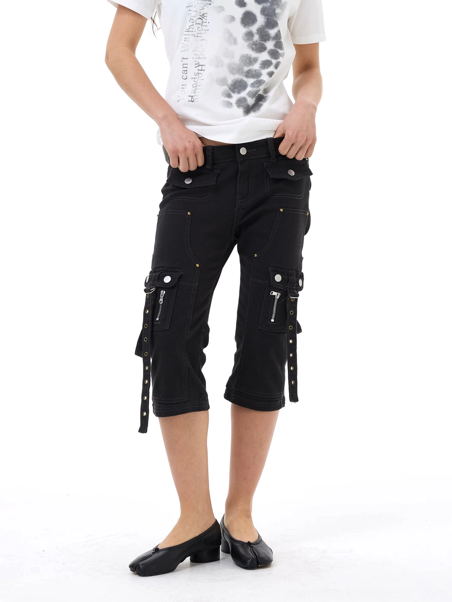 Front view of black strap cargo capri jeans styled with a printed white T-shirt and black flats, showing slim cropped fit, utility zipper pockets, metal rivets, and hanging side straps