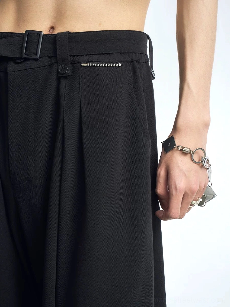 Close-up of black wide-leg streetwear pants waist showing adjustable belt buckle, front pleats, side zipper pocket, matte suiting fabric, and metallic hardware details