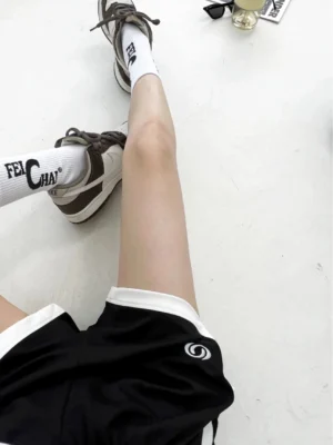 Close-up view of black streetwear track shorts showing white leg opening piping, lightweight knit cotton texture, and small circular front emblem
