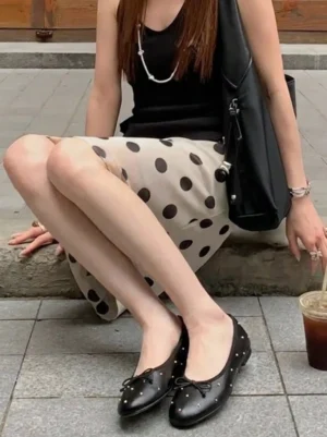 Woman wearing black studded ballet flats with a bow, polka dot midi skirt, black tote bag, and iced coffee