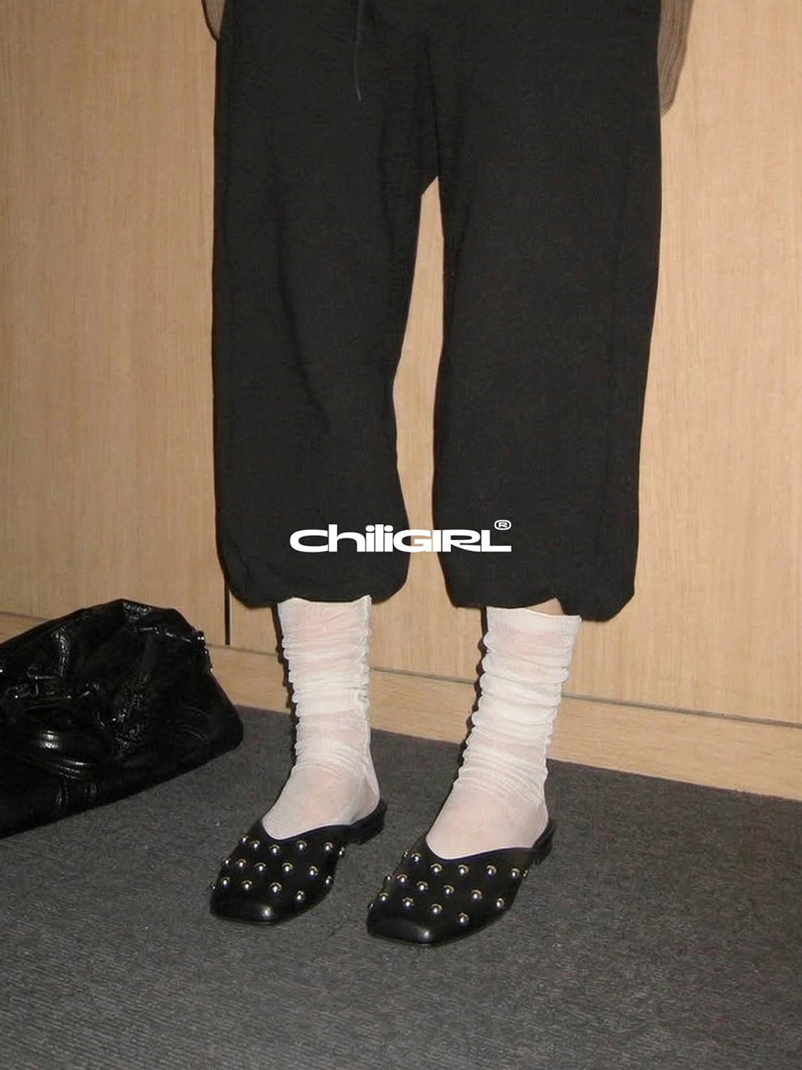 Black studded pointed-toe mule flats paired with slouchy white socks, cropped black lounge pants, and a minimalist leather tote