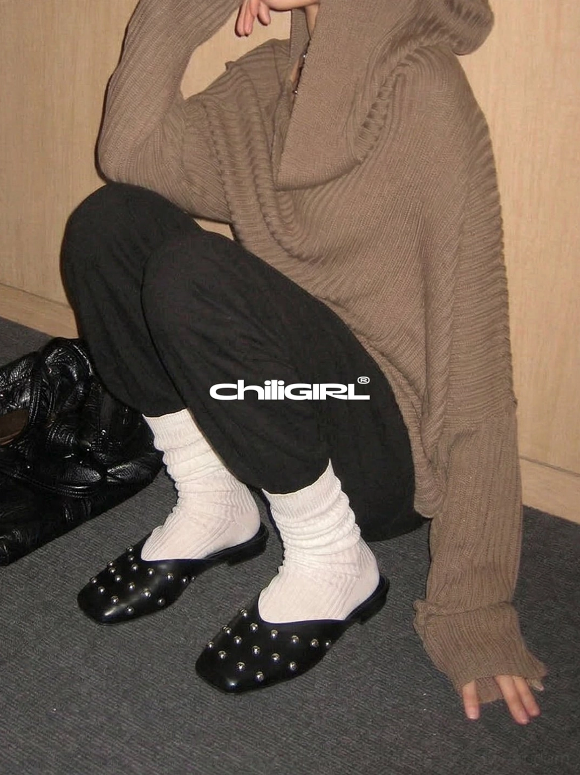 Black studded slip-on mule flats worn with slouchy white socks, cropped joggers, and an oversized brown knit hoodie