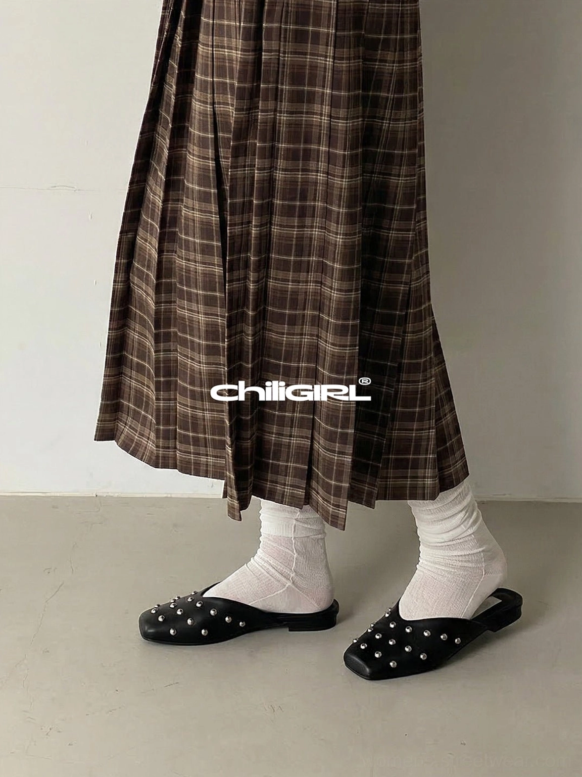 Silver-rivet black mule flats styled with white slouch socks and a brown plaid midi skirt for a retro preppy streetwear look
