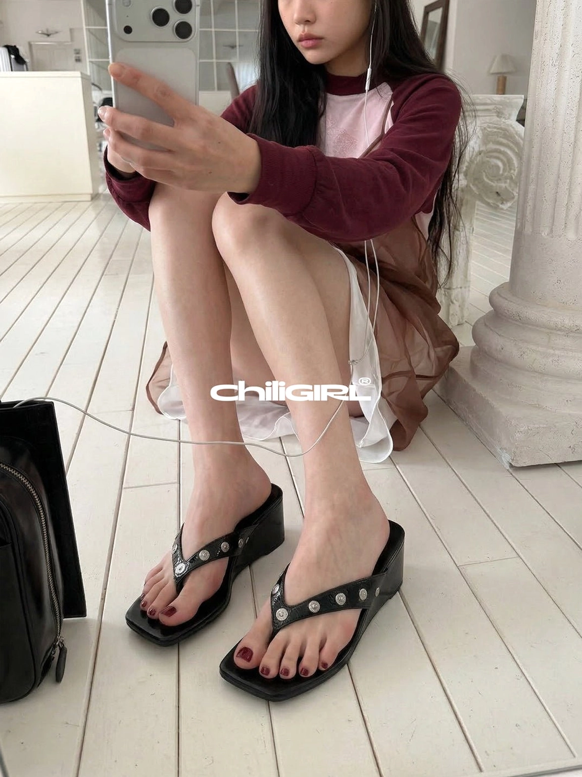 Woman seated on a wooden floor wearing black studded platform thong sandals with a modern square toe and urban streetwear styling