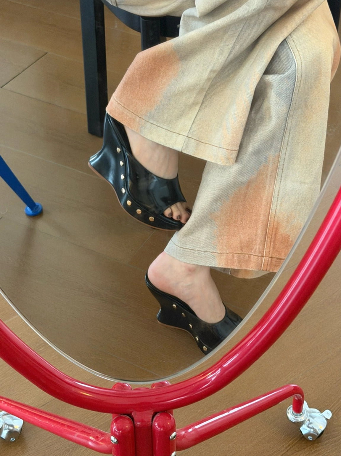 Low-angle mirror view of black studded platform wedge mules showing the dramatic wedge height, open-toe shape, rubber outsole, and metallic sole-edge details