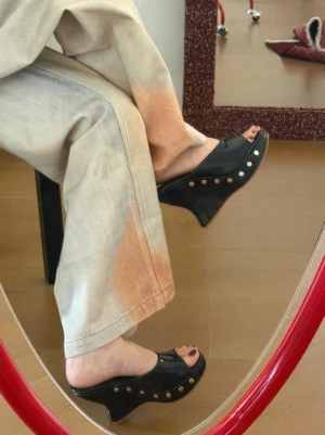 Black studded platform wedge mules worn with gradient wide-leg trousers in a mirror side view, highlighting the smooth upper and metallic rivet details