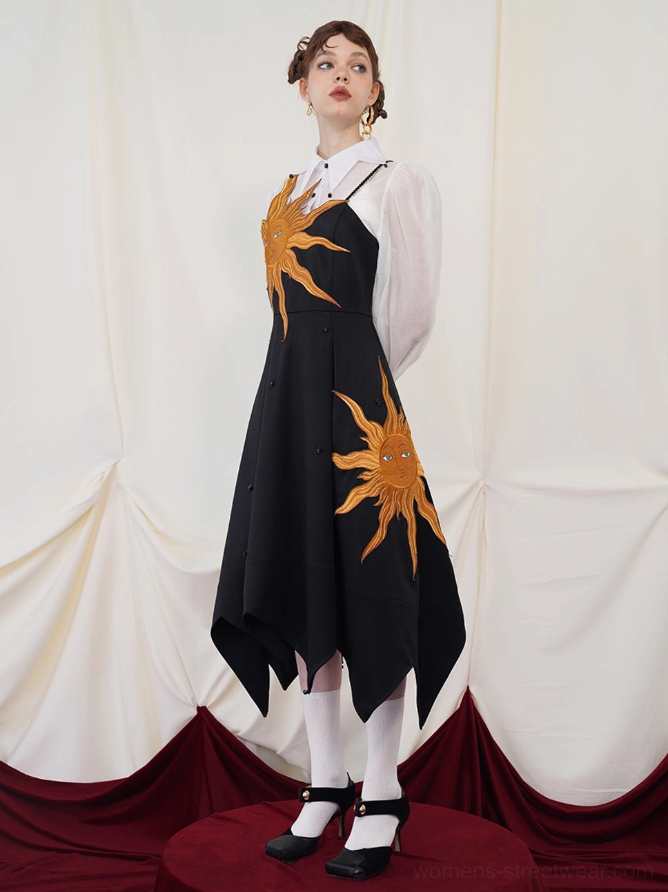 Side view of a black embroidered slip maxi dress showing two golden sun motifs and an irregular asymmetric handkerchief hem