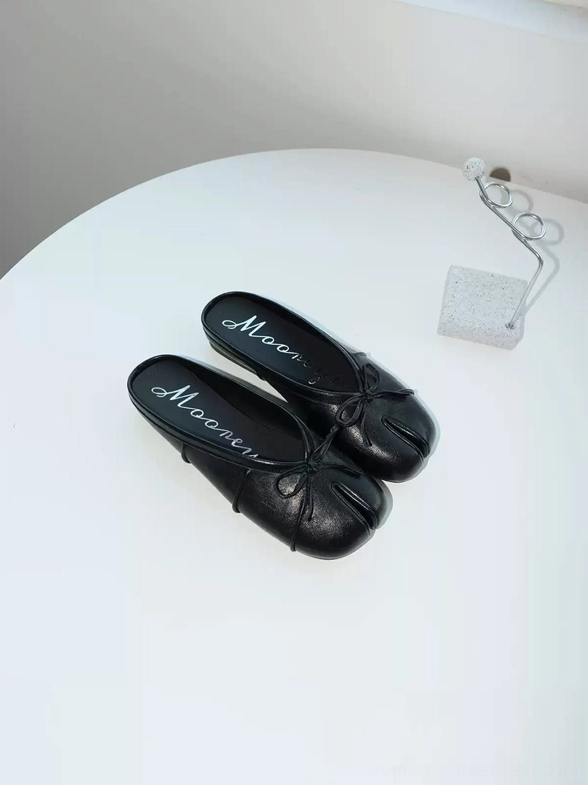 Pair of black tabi toe bow slip-on mules displayed on a white side table showing the smooth PU finish