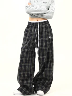Front left view of black tartan plaid wide-leg streetwear pants with elastic drawstring waist, white ruffled trim, embroidered emblem, and relaxed oversized fit styled with a cropped gray top and white sneakers