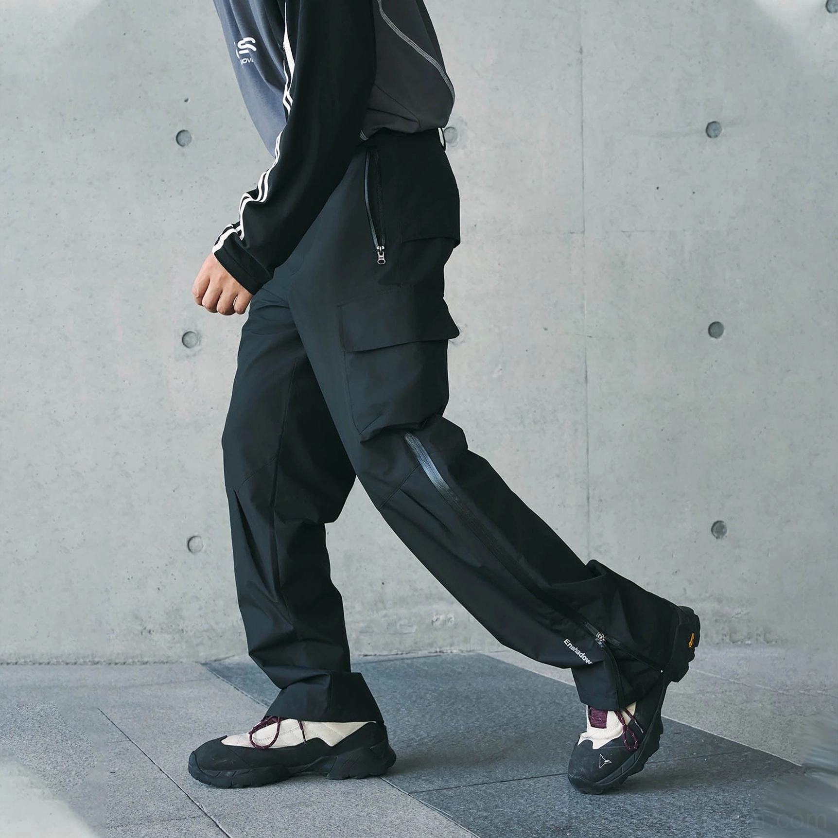 Black tech cargo waterproof pants worn in motion with a long side-leg zipper, thigh utility pocket, and relaxed urban techwear fit