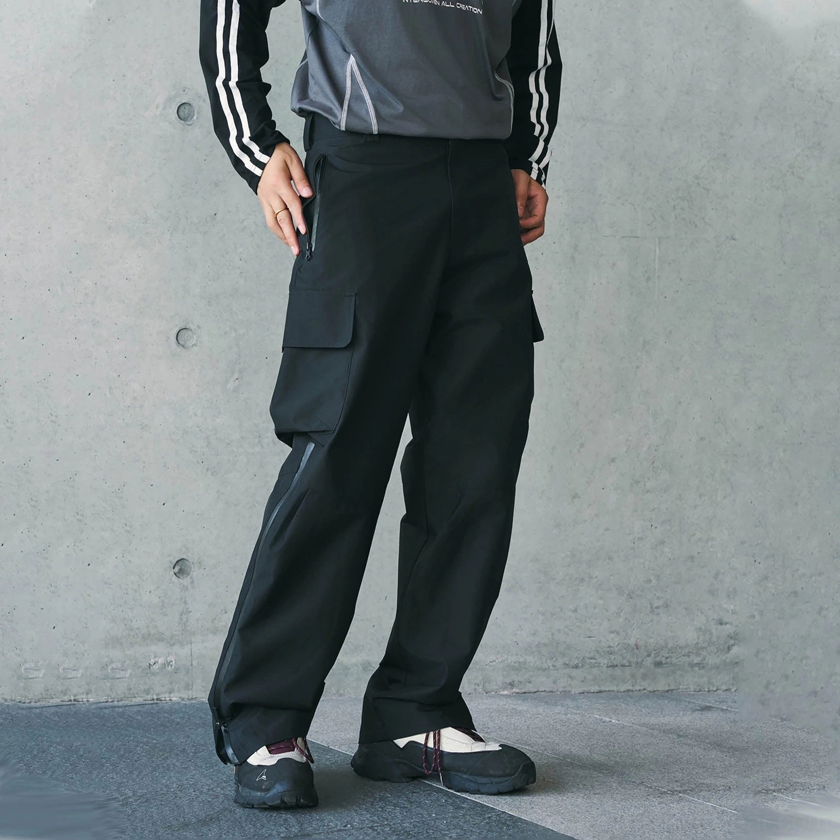 Black techwear waterproof cargo pants shown from a partial front side view with large cargo pockets, side-leg zippers, and a loose tactical silhouette