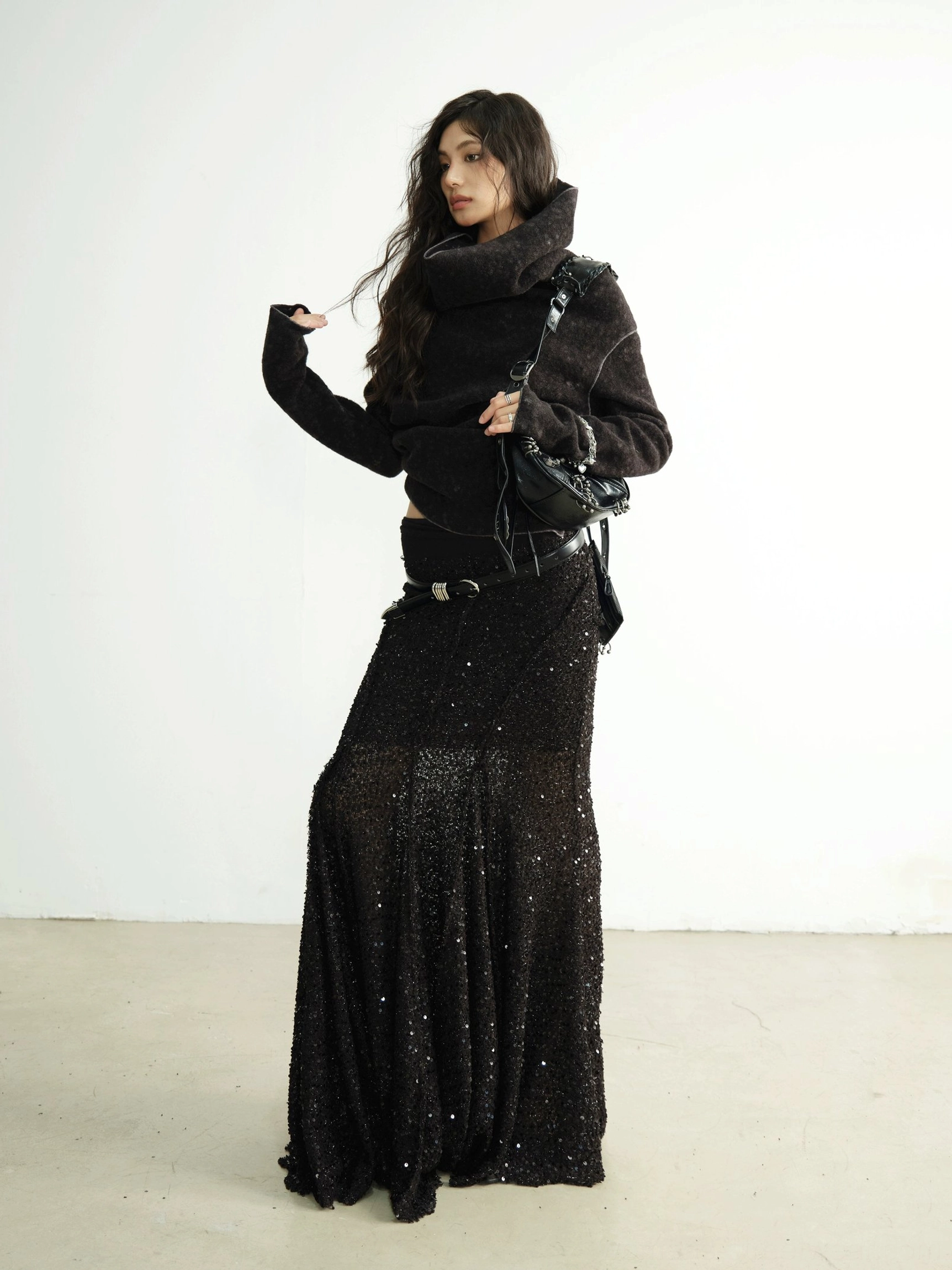 Side three-quarter view of a black textured cowl-neck knit cardigan with fitted silhouette, mermaid maxi skirt, and crossbody handbag