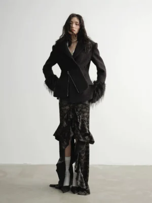 Full body three quarter view of a black textured faux fur cuff tailored coat with asymmetrical zipper worn with a ruffled slit maxi skirt and pointed heels