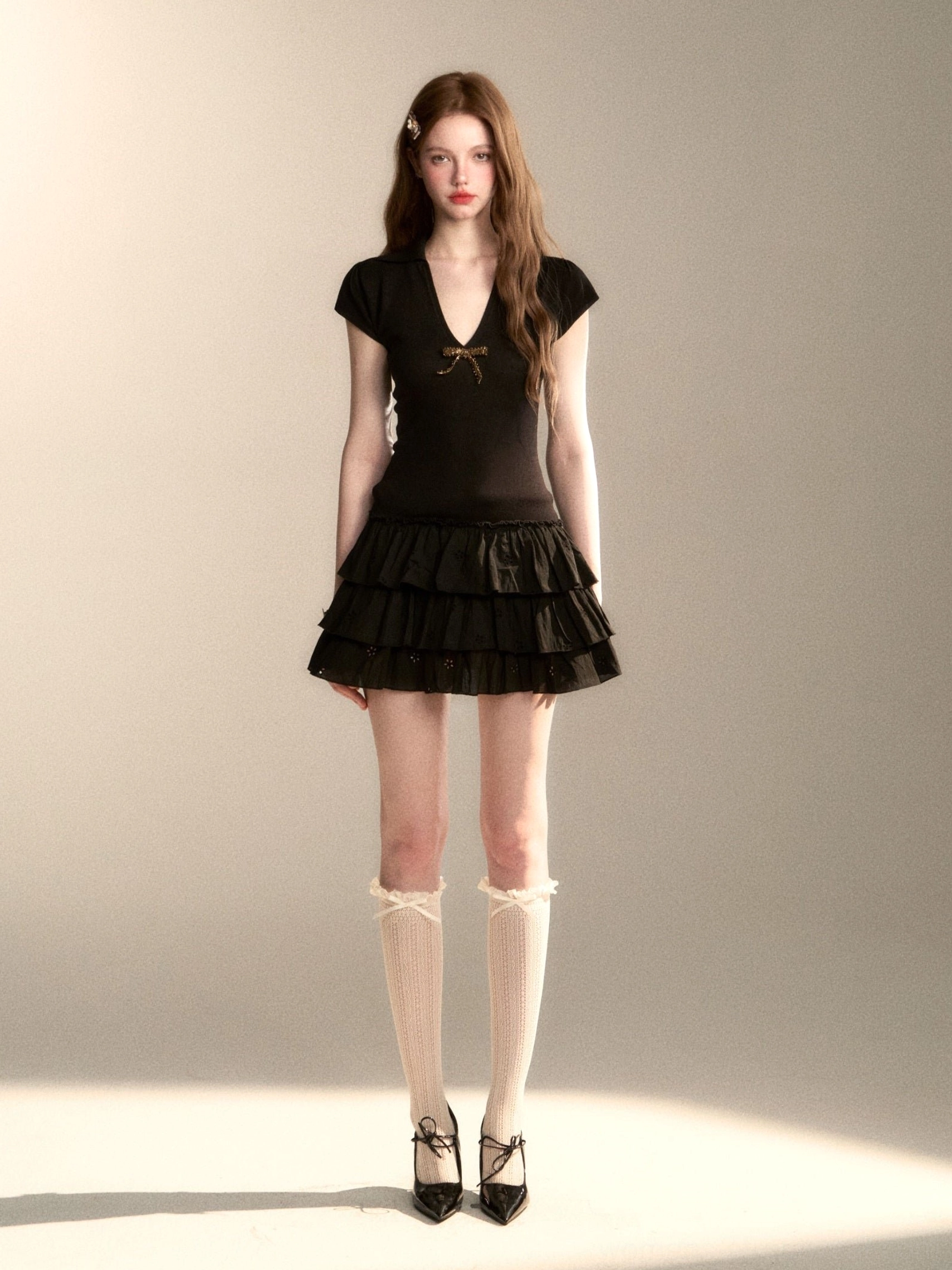 Full front studio look of black tiered ruffle mini dress with chest bow styled with lace knee socks and Mary Jane shoes
