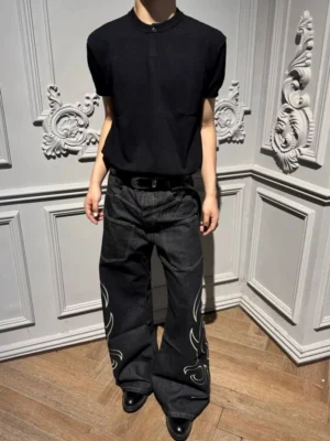 Front full-body view of black tribal flame wide leg jeans with white outer-leg embroidery, oversized pockets, stacked hems, and relaxed streetwear fit