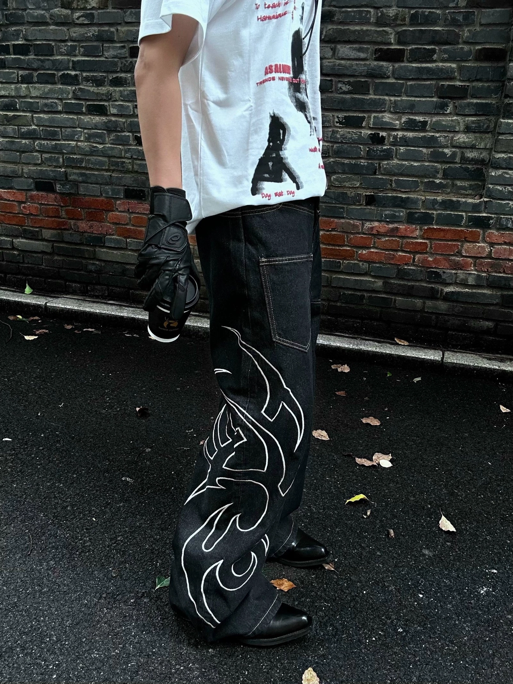 Side view of dark black wide leg denim jeans featuring white tribal tattoo flame embroidery, classic rear pocket, and oversized streetwear silhouette