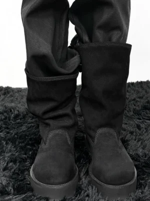 Model wearing black tumbled leather slouch mid-calf boots with loose dark trousers on a black textured rug, showing the thick platform outsole