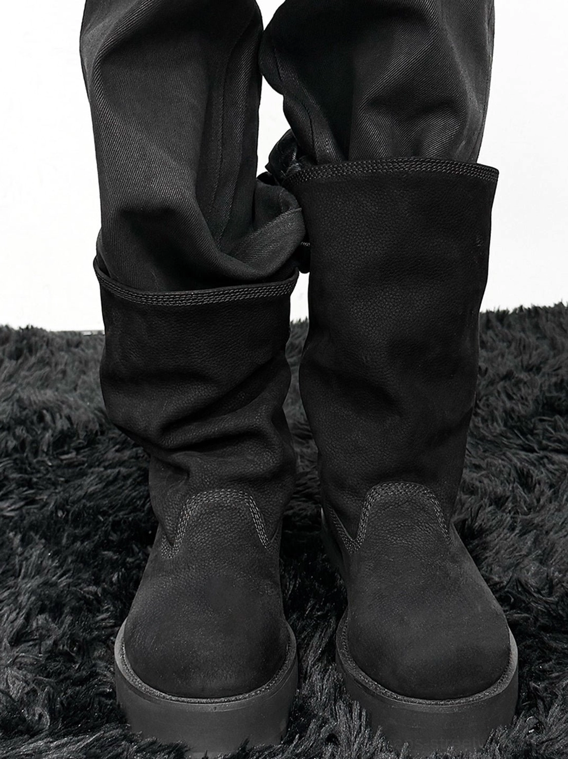 Model wearing black tumbled leather slouch mid-calf boots with loose dark trousers on a black textured rug, showing the thick platform outsole