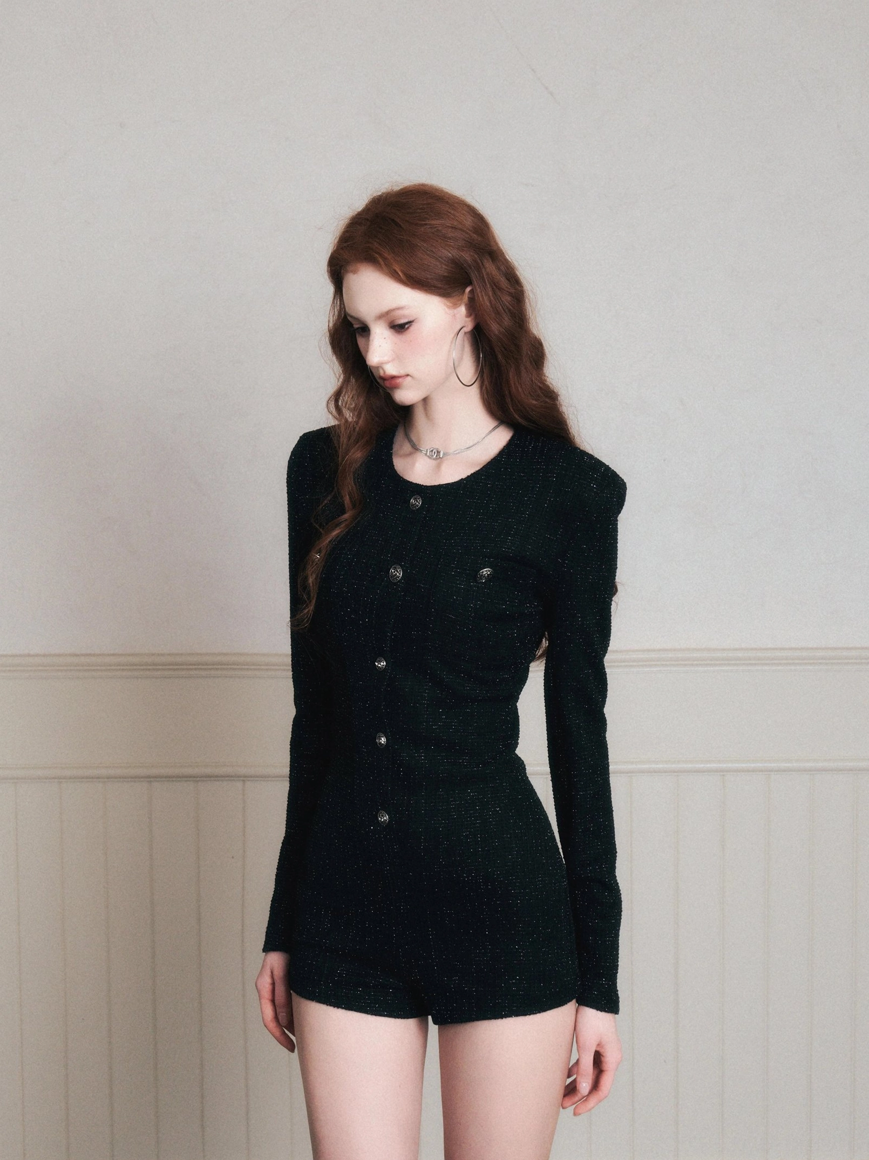 Front neutral full-body view of a black tweed romper with a minimalist fitted silhouette, short tailored shorts, long sleeves, and sleek textured finish