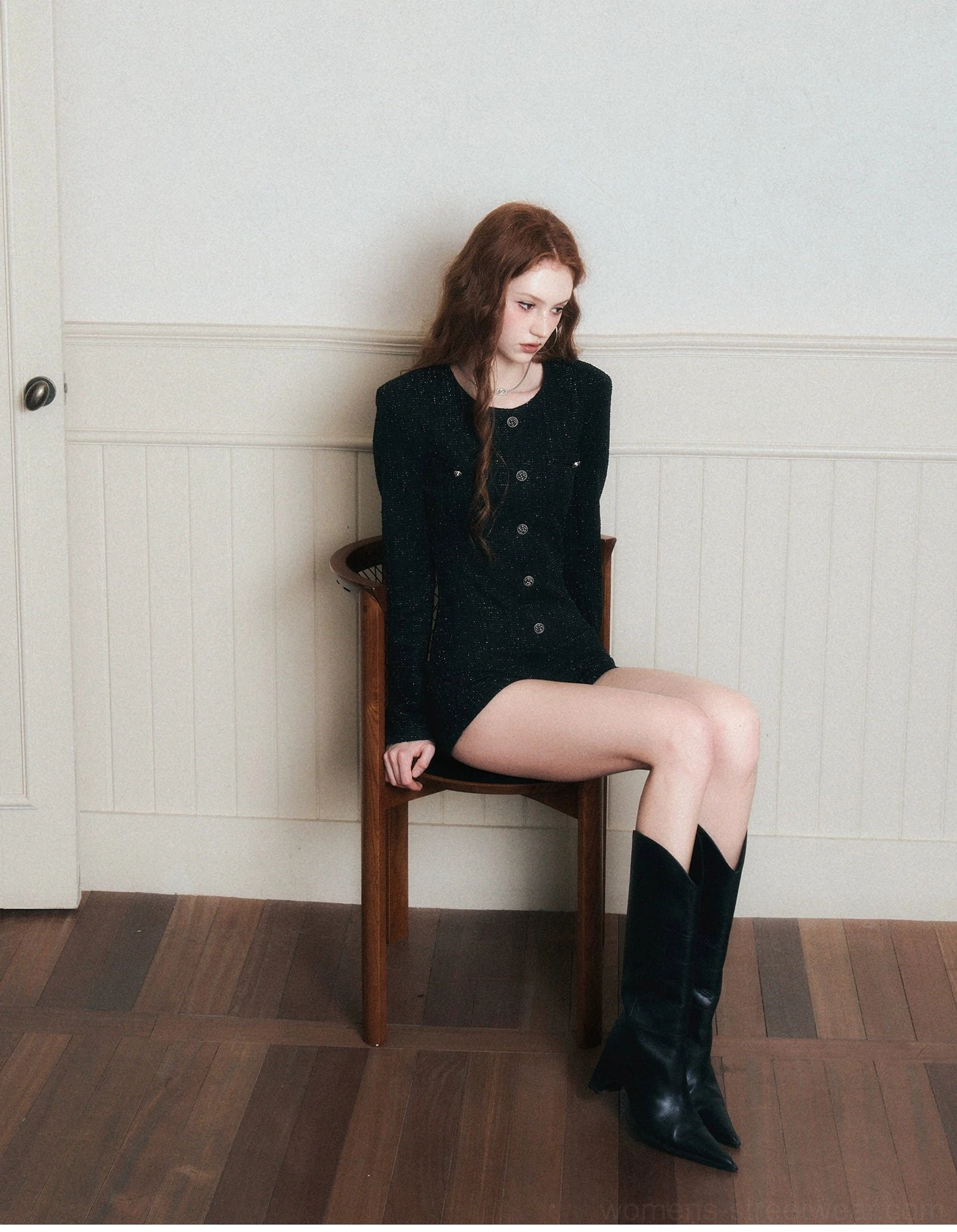 Seated front three-quarter view of a woman wearing a fitted black tweed romper with decorative buttons, tailored shorts, and knee-high leather boots