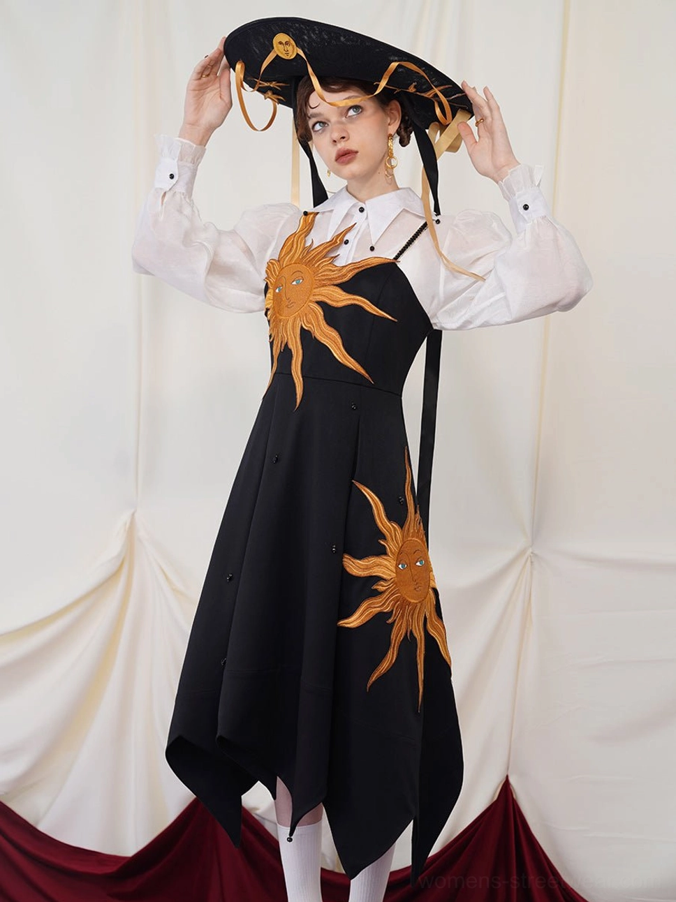 Front view of a black twilight sun embroidered asymmetric slip maxi dress styled with a sheer white blouse and wide brim hat