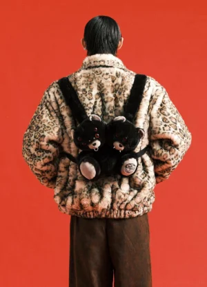 Male model wearing black twin evil bear faux fur mini backpack with a leopard print fuzzy coat against a red studio background