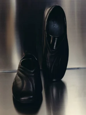 Pair of black low-profile leather slip-on shoes with curved wave stitching and glossy collar trim on metallic studio background
