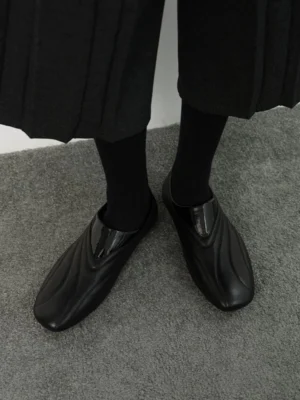 Black wave seam low-profile leather slip-on streetwear shoes worn with black socks and a midi skirt on indoor carpet