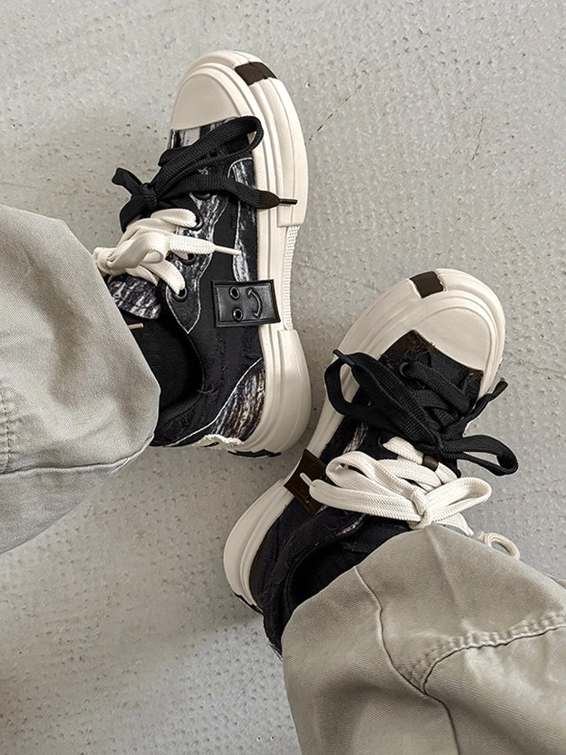 Top view of black and white distressed canvas platform sneakers with layered dual laces and gray cargo pants
