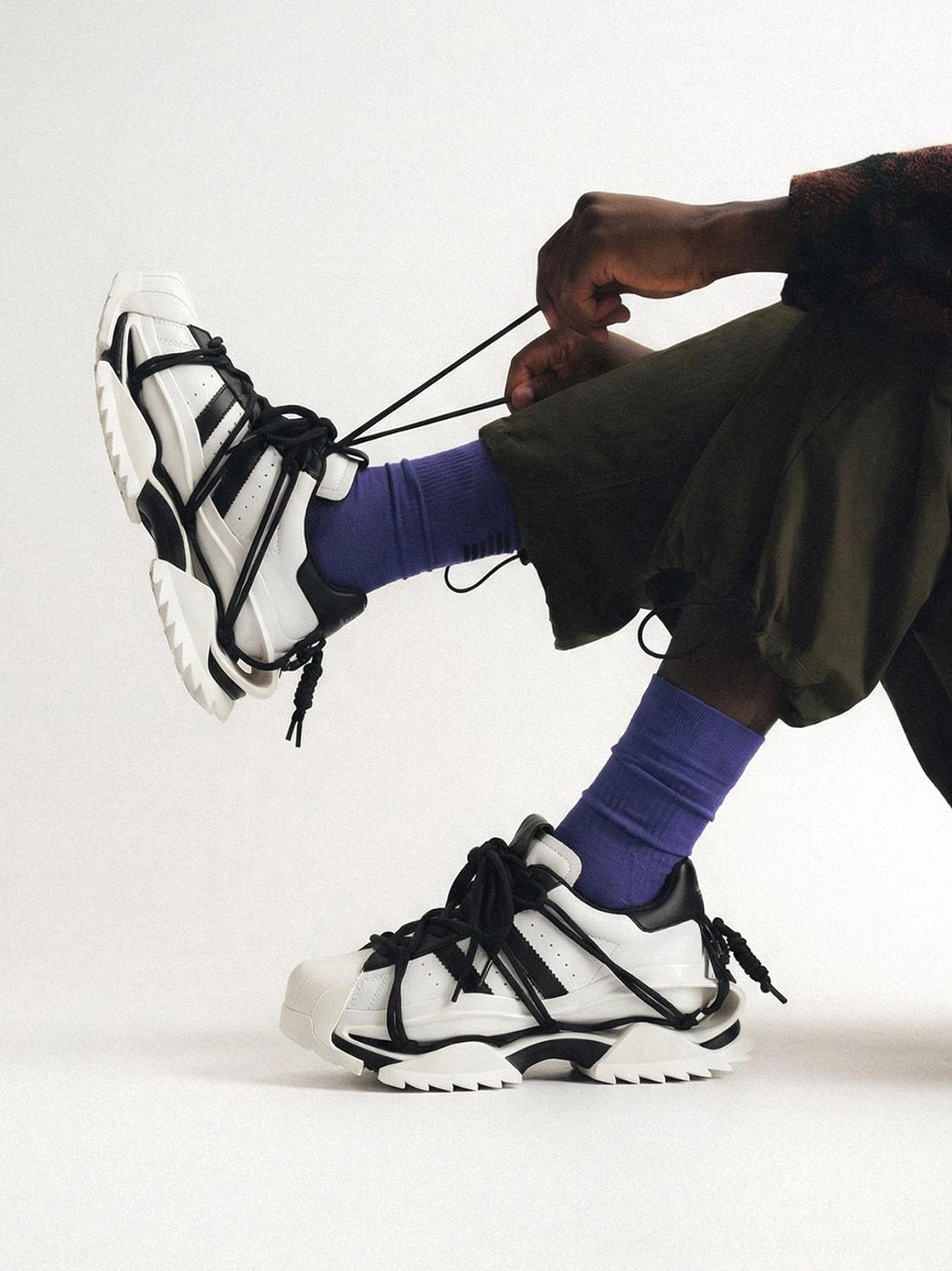 Low angle view of black and white chunky streetwear sneakers with layered straps and sculpted outsole worn with purple socks and dark green cargo shorts