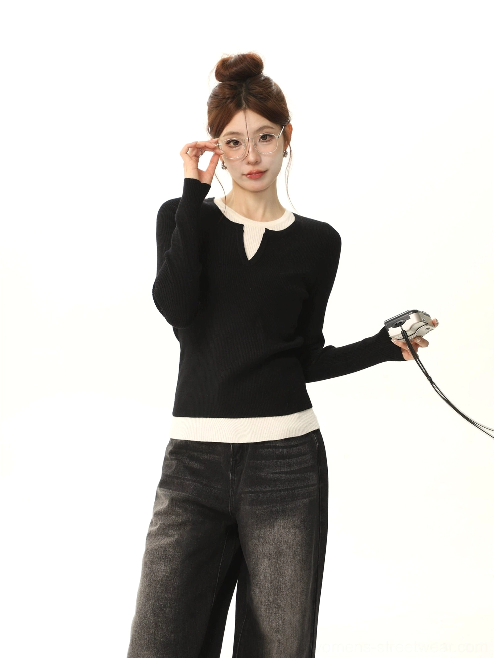 Half-body front view of a woman wearing a slim black and white color-block knit sweater with round neckline and white contrast trim, styled with clear-frame glasses and a vintage camera accessory
