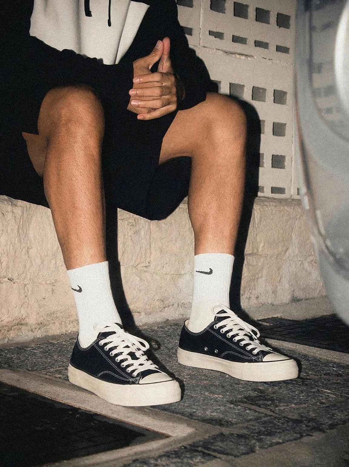 Person wearing black and white low-top canvas platform sneakers with cork midsole, sitting outdoors in casual streetwear outfit with black shorts and socks